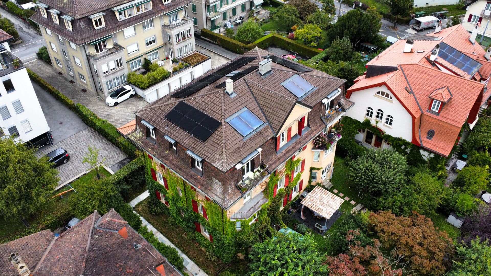 Aerial view of a residential area with solar panels on rooftops, surrounded by greenery and neighboring buildings.